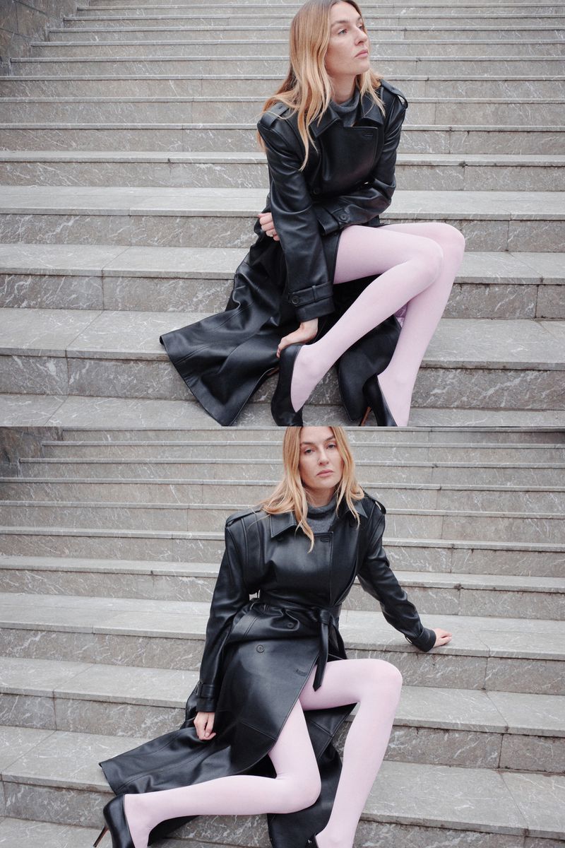 Blonde woman in a long black leather trench coat and pink tights poses on grey stone steps with black high heels, fashion portrait.