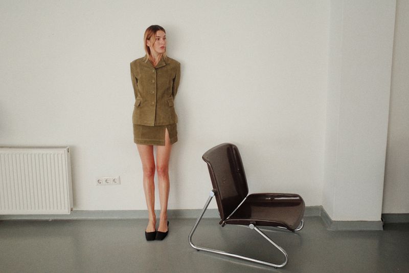 A woman posing in a beige skirt suit next to a brown chair in an indoor setting with white walls and gray floor and a radiator.