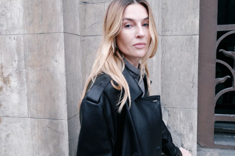 A blonde woman in a black leather trench coat stands in front of a stone wall with light filtering through on an overcast day.