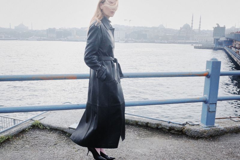 Woman in long black leather coat and heels stands on waterfront, overlooking a misty cityscape with mosques and waterways in background.