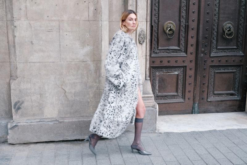 Blonde woman in an animal print coat walking past a stone wall and an old wooden door on a city street wearing tights and heels.