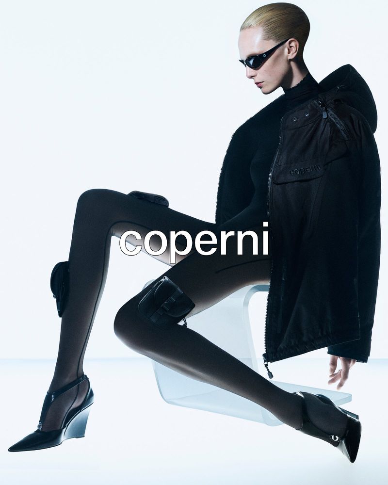 Fashion shot of a woman with sunglasses in a black jacket and stockings, seated in a clear chair against a bright studio backdrop.