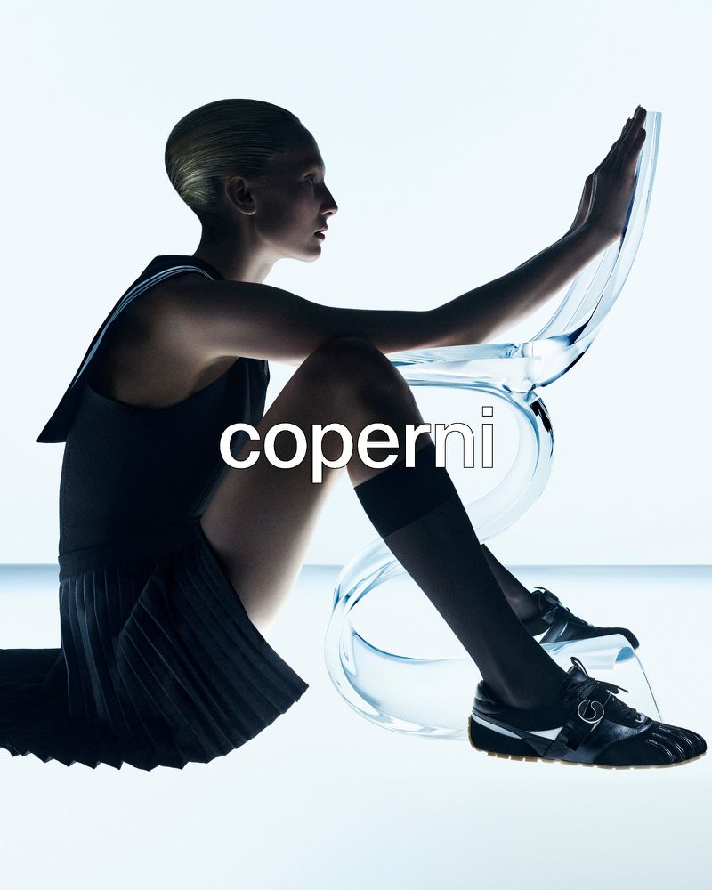 A model wearing a dark outfit with a clear chair on a white background in a studio shot, showcasing fashion and beauty.