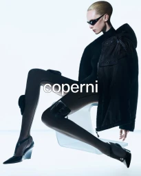 Model Alex Consani wearing dark sunglasses and a black hooded jacket against a white studio background for Coperni
