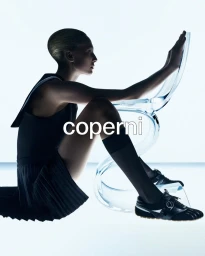 Model Alex Consani posing seated on the floor touching a transparent chair for Coperni F/W 2025 Campaign