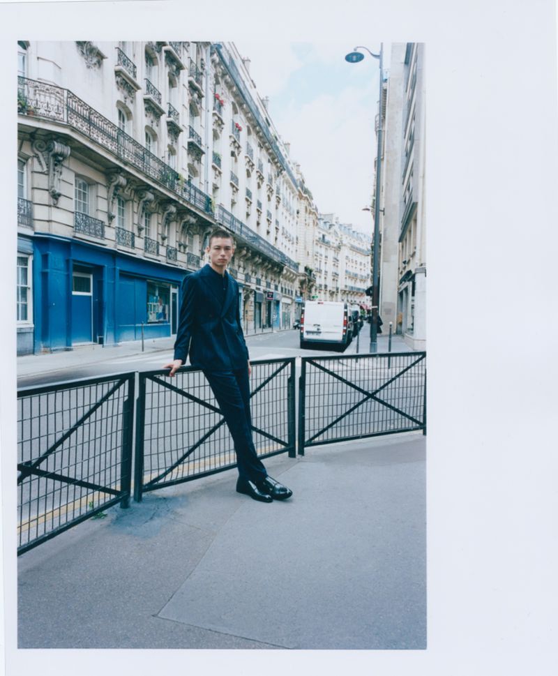 Young man in a dark suit leans against a metal railing on a city street with classic European architecture, van in background.