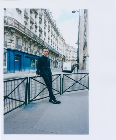 Young man in a dark suit leans against a metal railing on a city street with classic European architecture, van in background.
