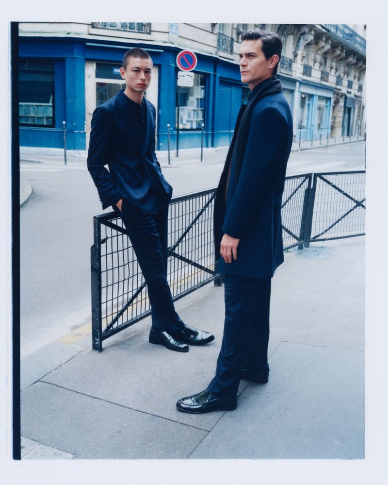 Two men in dark suits stand on a city street near a blue building, looking away in a fashion editorial style photograph on a gray day.