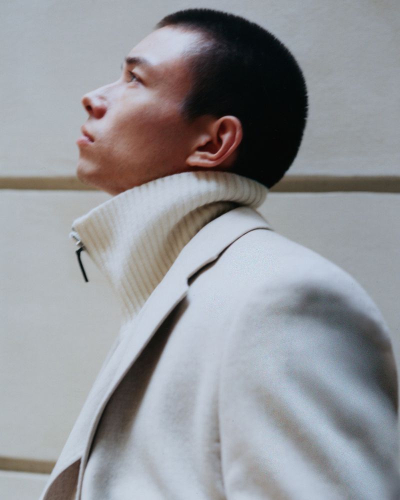Man in a beige suit and turtleneck sweater looks upward in a close-up shot, with a clean neutral background, and a shallow depth of field.