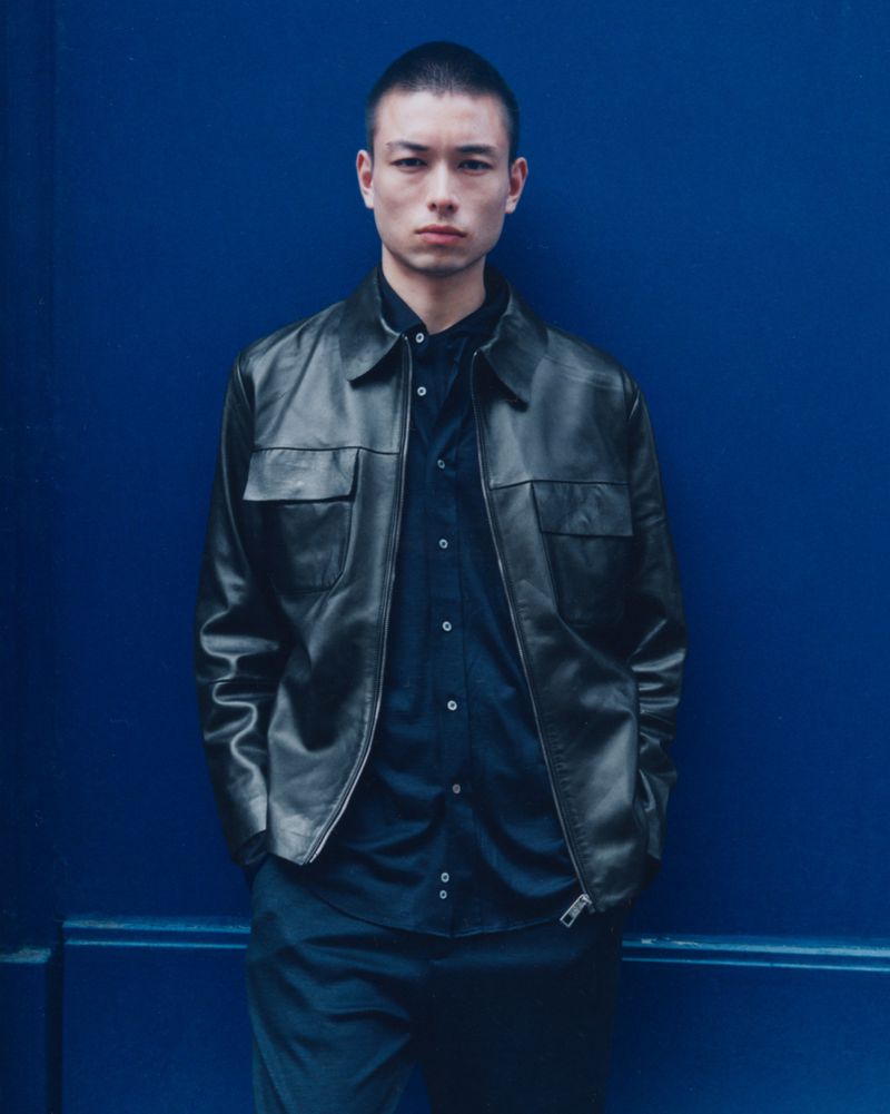 Portrait of a young adult male wearing a black leather jacket and black collared shirt against a solid royal blue backdrop.
