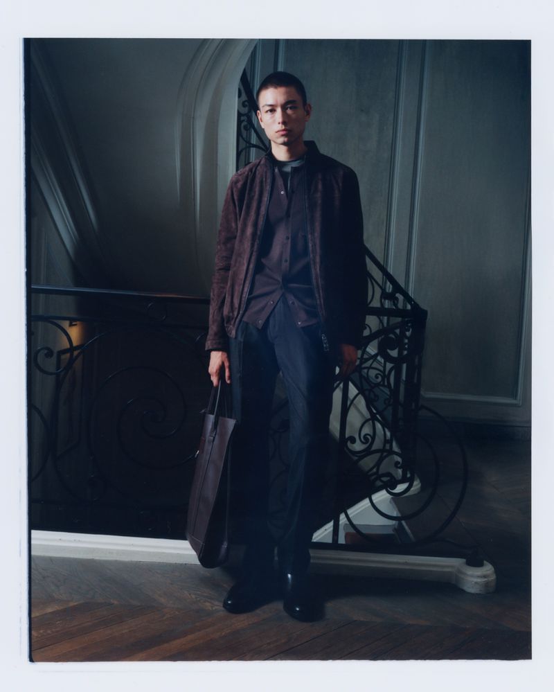 Fashionable adult male poses indoors wearing casual jacket, shirt, and dark pants, holding a tote bag with a stairway background.