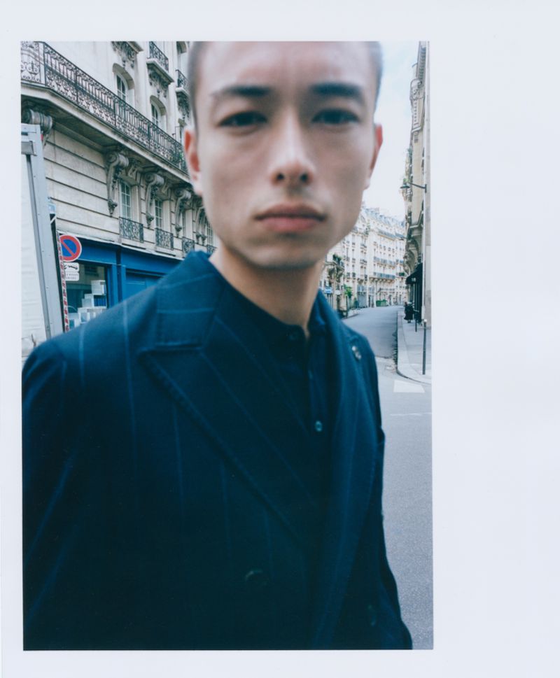 Portrait of an Asian man in a dark blue suit on a European city street with historical buildings in the background on an overcast day.