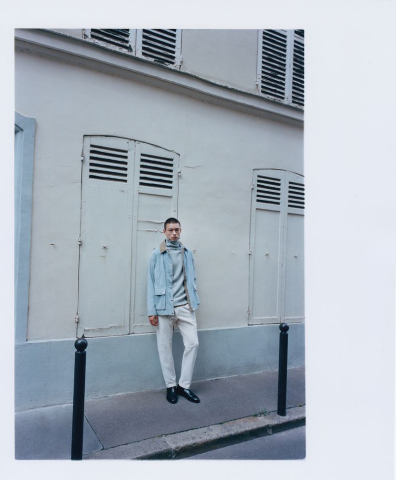 A young adult male in a light blue jacket poses in front of an off-white building with gray shutters on a city street in an outdoor shot.