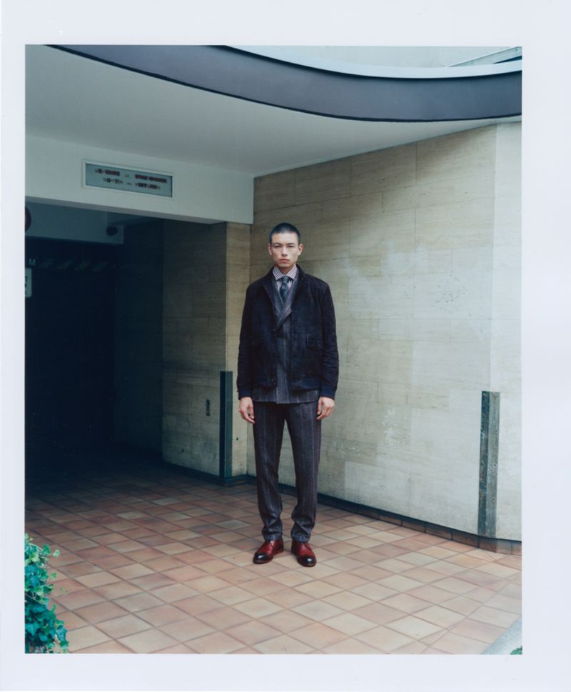 Full shot of a young adult male standing in a suit in front of a textured wall and opening to a dark area, with modern architecture.