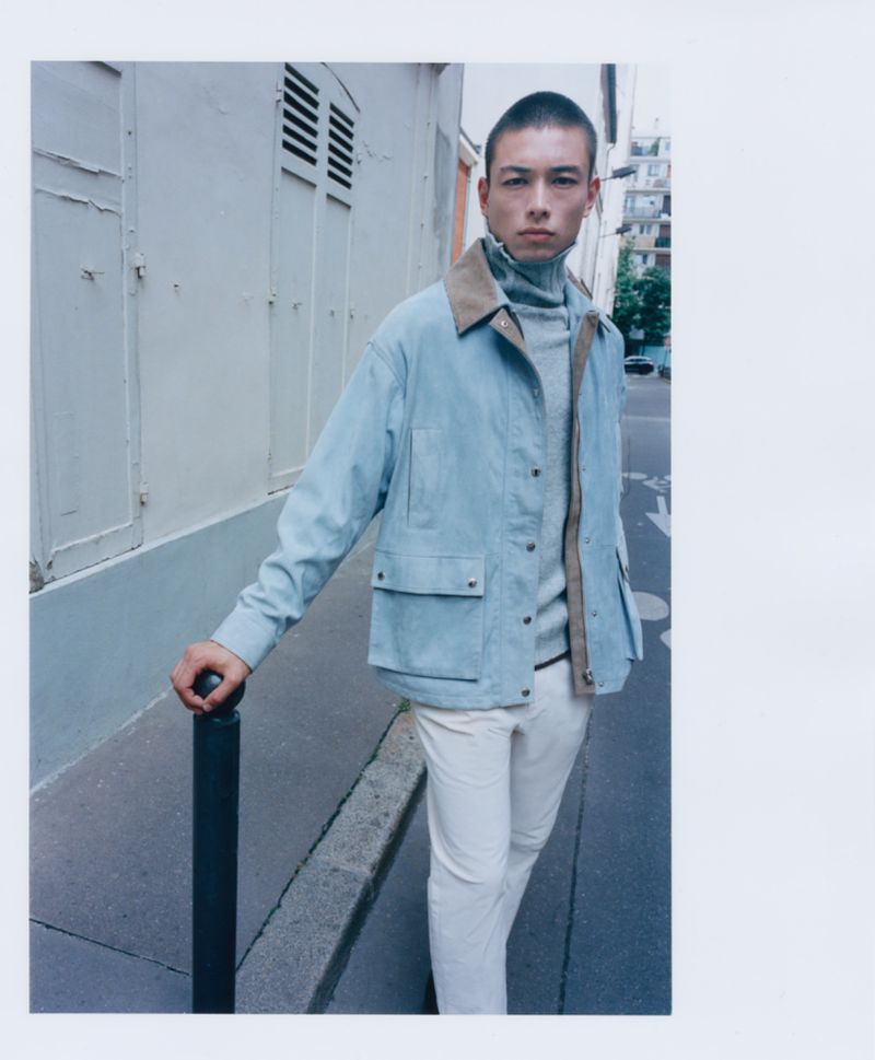 Man in light blue jacket, gray turtleneck, and white pants poses on city street with light-colored buildings in the background.