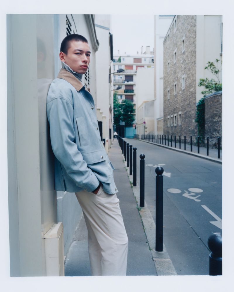 Young adult male model in a light blue jacket leaning against a building on a city street with urban architecture in the background.