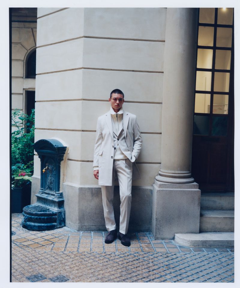 Adult male fashion model posing in an off-white suit with a column and water fountain as a background, captured in a medium shot.