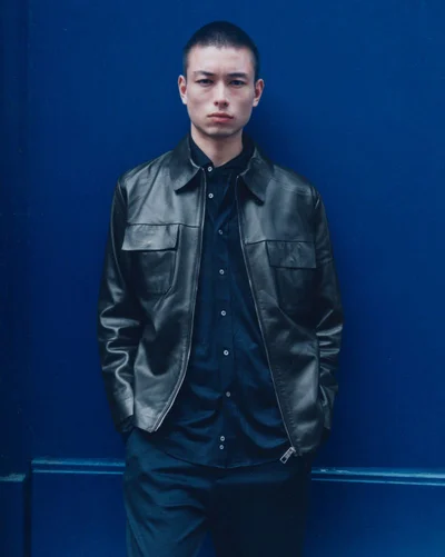 Model Nicolas Yoshi wearing an open black leather jacket over a navy buttoned shirt against a solid blue background.