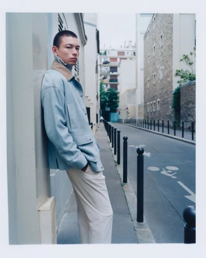 Model Nicolas Yoshi wearing a light blue jacket with a tan collar and cream trousers, leaning against a building on a city street.