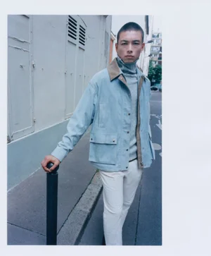 Model Nicolas Yoshi wearing a light blue jacket and white trousers, leaning on a street bollard in an urban setting.