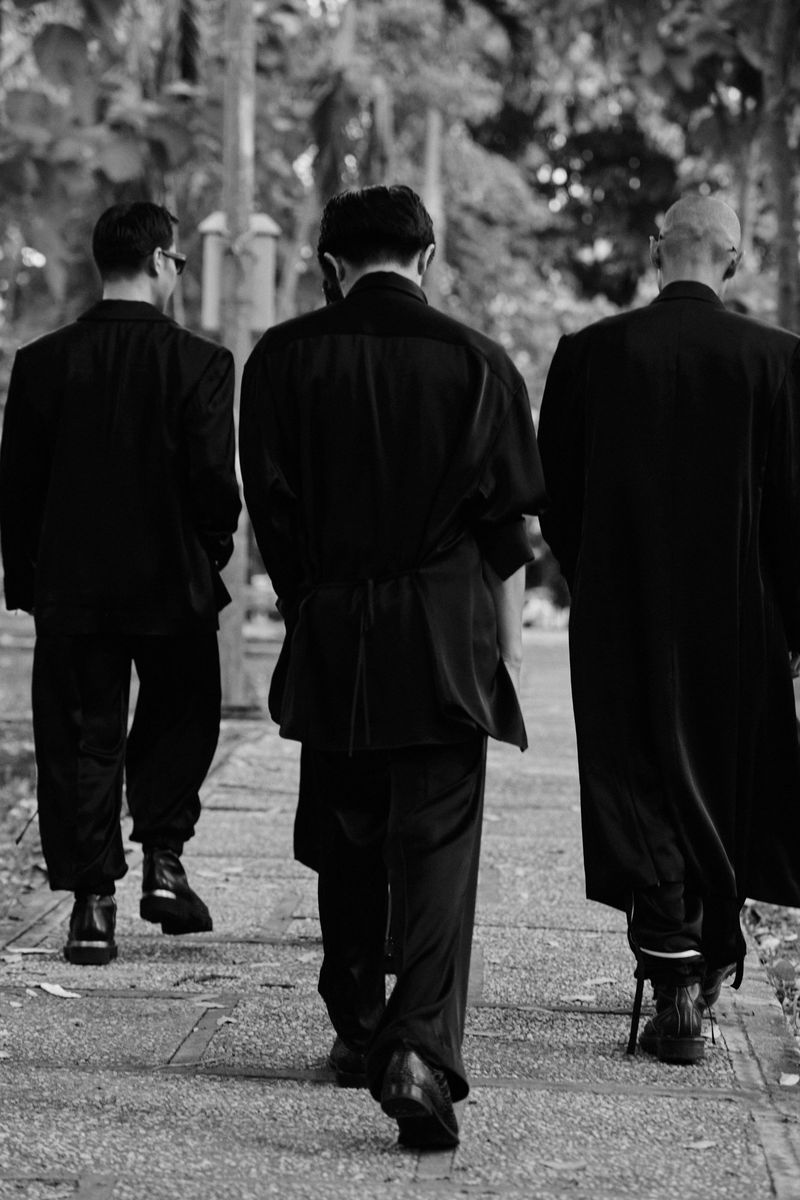 Three adult men in dark suits walk away on a sidewalk lined with trees, captured in black and white with a shallow depth of field.
