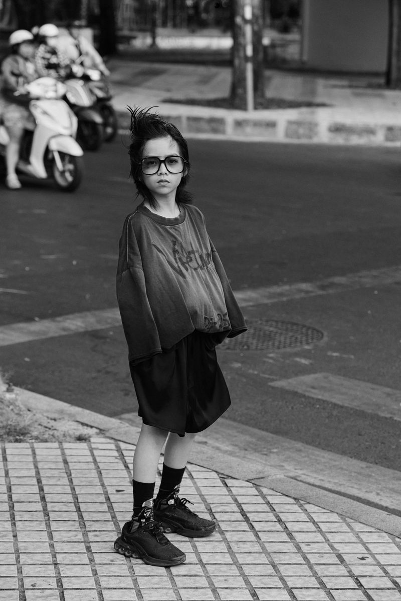 Child in oversized T-shirt and shorts stands on a city street with scooters blurred in the background, captured in black and white.