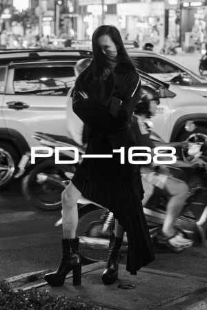 A person poses on a street corner at night, wearing a dark jacket, skirt, and platform boots, standing in front of traffic.