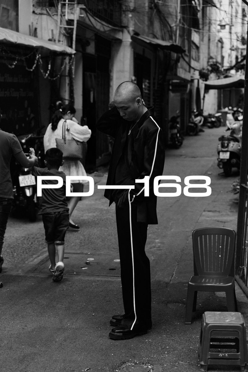 Monochrome street scene featuring a slender adult male in a dark suit standing near a child and adults in a bustling urban alleyway.