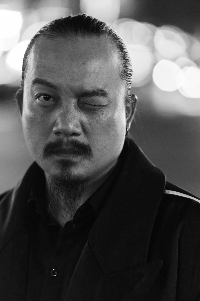 Monochrome portrait of an adult male winking at the camera in an urban setting, wearing a dark coat, with blurred city lights in background.