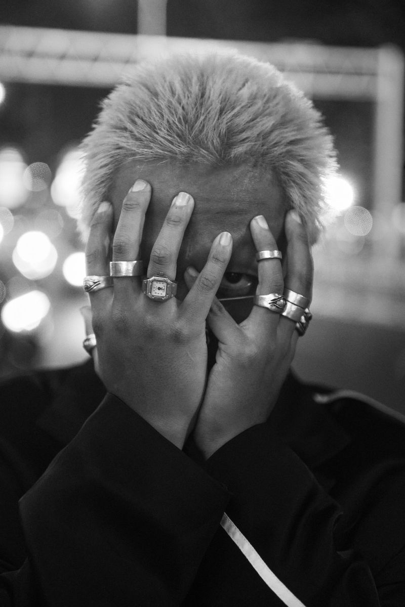 Black and white portrait of a person with dyed blonde hair covering their face with hands adorned with multiple rings, dark clothing.