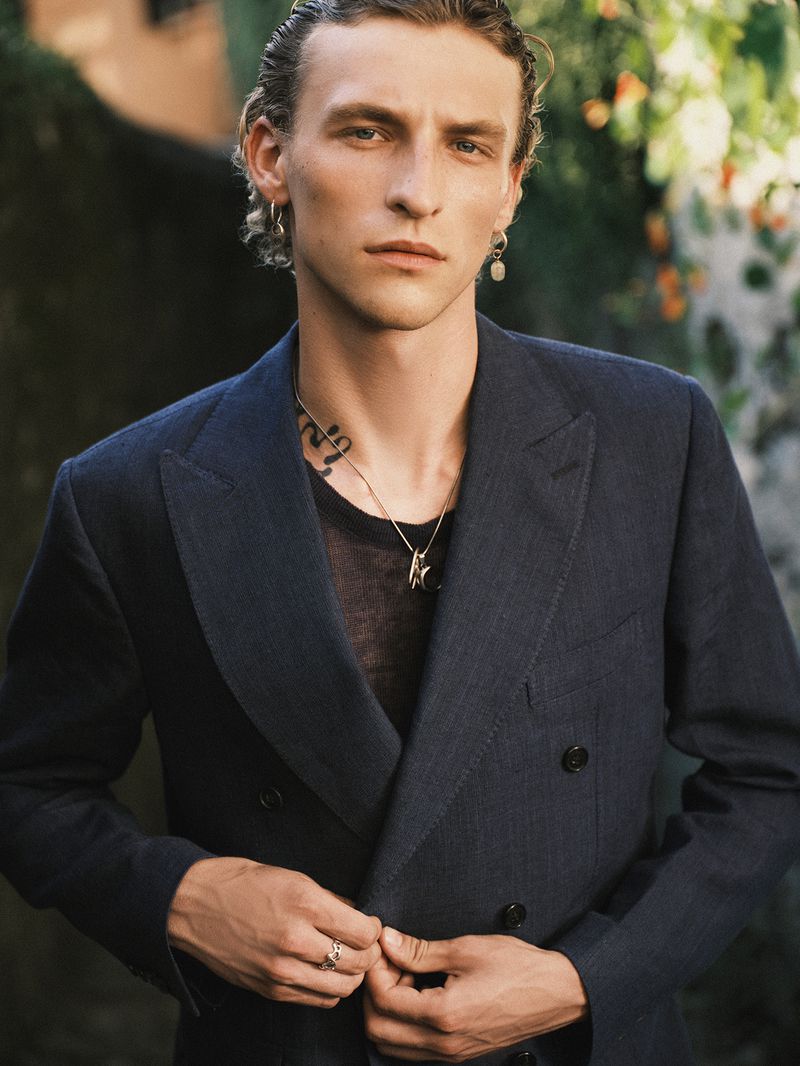 Portrait of an adult male wearing a navy blazer, t-shirt and jewelry in front of a lush green background, medium shot, eye-level angle.