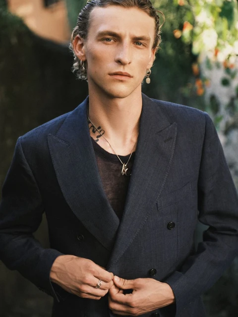 Portrait of an adult male wearing a navy blazer, t-shirt and jewelry in front of a lush green background, medium shot, eye-level angle.