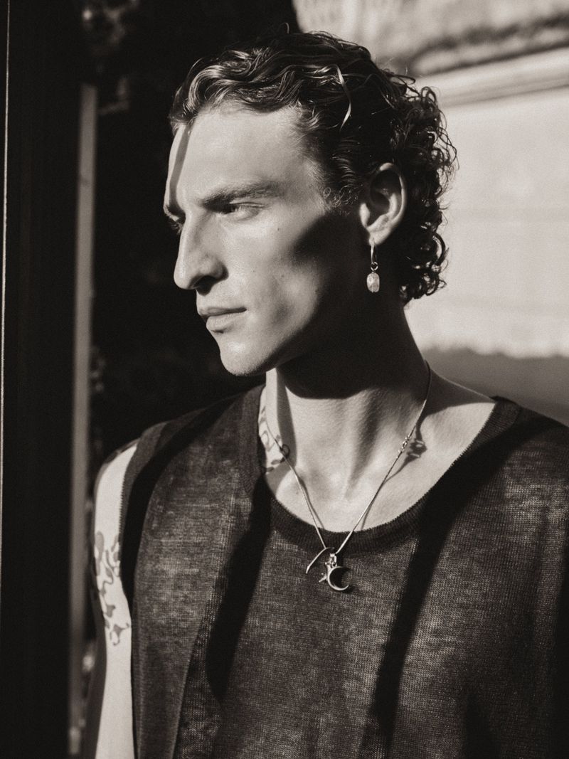 Portrait of a male model with dark curly hair and earrings. He is wearing a black tank top with a necklace. Strong shadows.