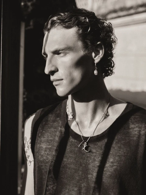 Portrait of a male model with dark curly hair and earrings. He is wearing a black tank top with a necklace. Strong shadows.