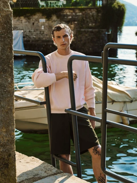 Adult male poses on stairway near lake wearing pink sweatshirt and dark shorts with boat and building in background on sunny day.