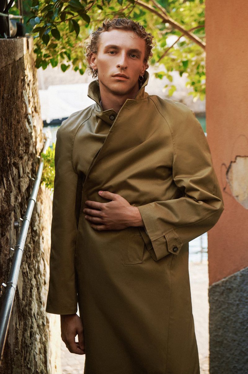 Man in trench coat poses in outdoor setting against old walls and greenery. Fashion portrait with natural light and neutral palette.