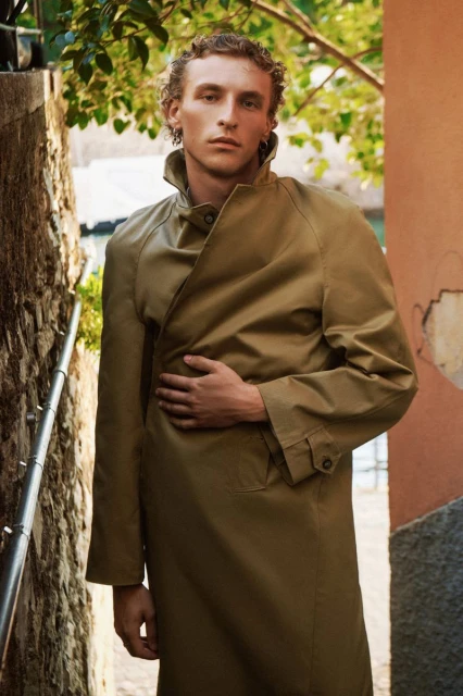 Man in trench coat poses in outdoor setting against old walls and greenery. Fashion portrait with natural light and neutral palette.