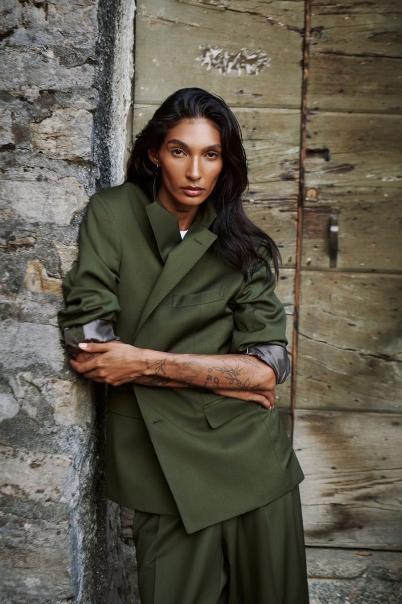 Adult female model poses in an olive-green suit, arms crossed, leaning against a stone wall and weathered wooden door in an outdoor setting.