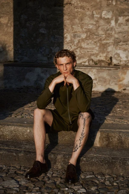 Young adult male in olive green shorts suit and leather shoes sits on stone steps with hands clasped under chin in afternoon sun.