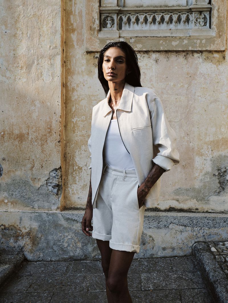 Fashionable adult woman poses in a matching light-colored jacket and shorts in front of a textured building wall in natural light.