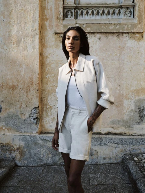Fashionable adult woman poses in a matching light-colored jacket and shorts in front of a textured building wall in natural light.