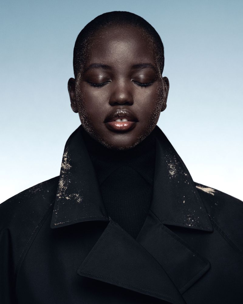 Portrait of a young Black woman in a dark trench coat with white sand-like particles on her face and shoulders against a light blue background.