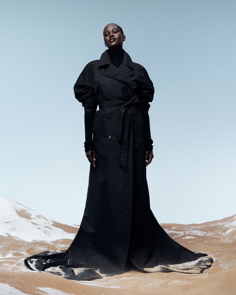 Elegant woman in black trench coat stands on a textured tan surface against a bright blue sky, showcasing fashion and style in a desert-like setting.