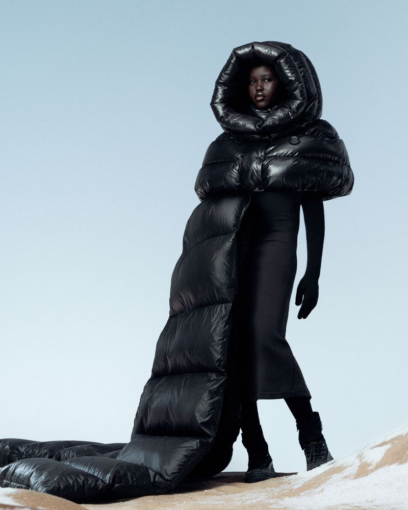 Fashion portrait of a Black model wearing an oversized black puffer coat with a hood against a gradient blue background in a studio.