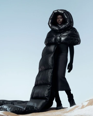 Fashion portrait of a Black model wearing an oversized black puffer coat with a hood against a gradient blue background in a studio.