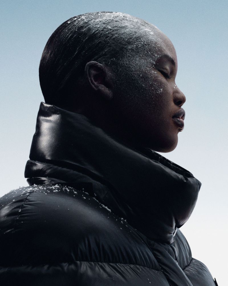 Profile of a woman with closed eyes, dusted with white powder, wearing a glossy black puffer jacket against a light blue gradient backdrop.