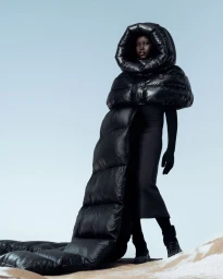 Model Adut Akech wearing an oversized sculptural black hooded puffer cape with a long train on a snowy slope under a clear sky.