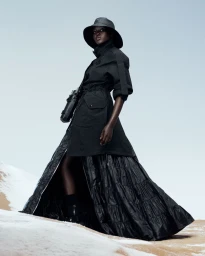 Model Adut Akech wearing a black belted coat and bucket hat standing on a snowy desert dune against a pale blue sky