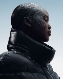 Adut Akech in profile wearing a black Moncler puffer jacket with frost dusted across her face against a pale gradient backdrop.