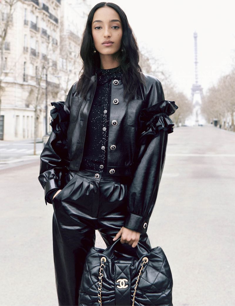 Fashion shot of a woman in a black leather jacket, black pants, and a Chanel purse, standing on a Paris street with the Eiffel Tower behind.